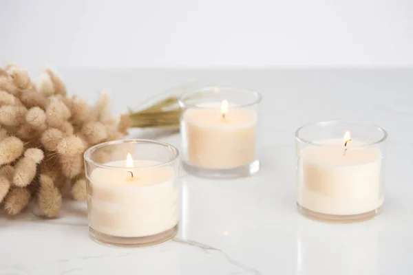 Selective focus of fluffy bunny tail grass and burning white candles in glass glowing on marble white surface — Stock Photo