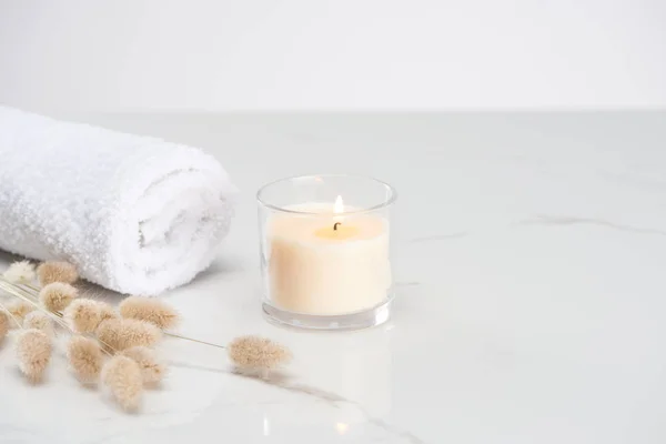 Fluffy bunny tail grass near burning white candle in glass and rolled towel on marble white surface — Stock Photo