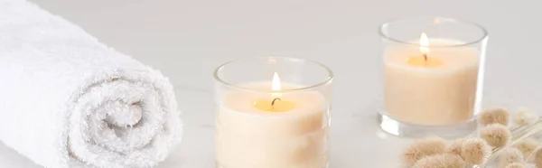 Fluffy bunny tail grass near burning white candles in glass and rolled towel on marble white surface, panoramic shot — Stock Photo