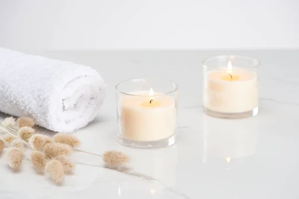Fluffy bunny tail grass near burning white candles in glass and rolled towel on marble white surface — Stock Photo