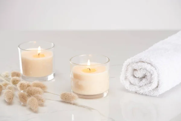 Flauschiges Hasenschwanzgras in der Nähe brennender weißer Kerzen im Glas und gerolltem Handtuch auf marmorweißer Oberfläche — Stockfoto