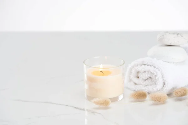 Fluffy bunny tail grass near burning white candle in glass and rolled towel with stones on marble white surface — Stock Photo