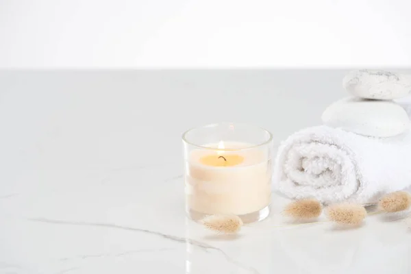 Fluffy bunny tail grass near burning white candle in glass and rolled towel with stones on marble white surface — Stock Photo