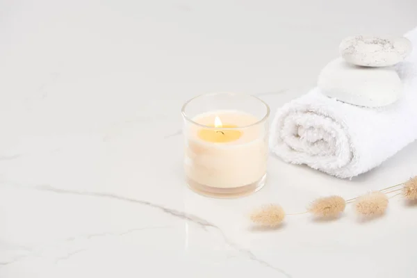 Flauschiges Hasenschwanzgras in der Nähe brennender weißer Kerze im Glas und gerolltem Handtuch mit Steinen auf marmorweißer Oberfläche — Stockfoto