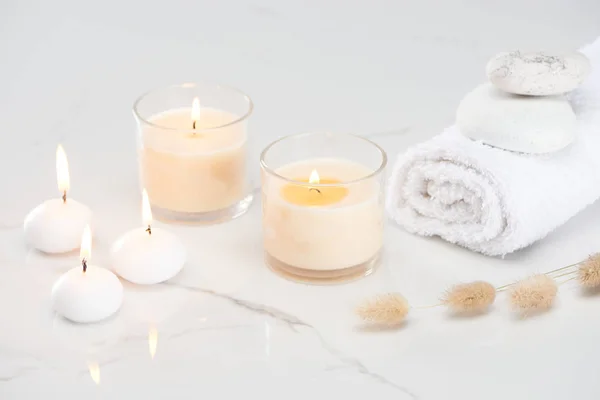 Fluffy bunny tail grass near burning white candles in glass and rolled towel with stones on marble white surface — Stock Photo