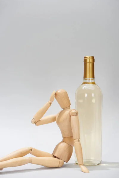 Muñeca de madera con la mano por la cabeza al lado de la botella de vino sobre fondo gris - foto de stock
