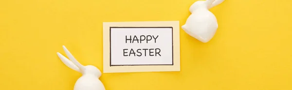 Top view of greeting card with happy Easter lettering near Easter white bunnies on yellow colorful background, panoramic shot — Stock Photo