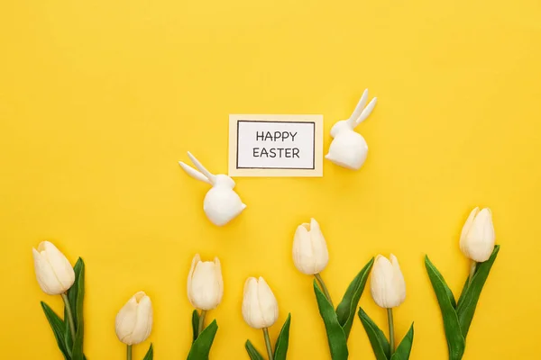 Top view of tulips, greeting card with happy Easter lettering near Easter white bunnies on yellow colorful background — Stock Photo