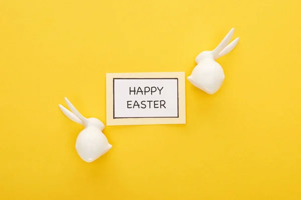 Top view of greeting card with happy Easter lettering near Easter white bunnies on yellow colorful background — Stock Photo