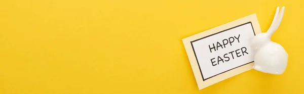Vista superior de la tarjeta de felicitación con letras de Pascua feliz cerca de conejito blanco de Pascua sobre fondo colorido amarillo, tiro panorámico - foto de stock