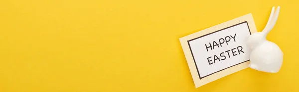 Vista superior de la tarjeta de felicitación con letras de Pascua feliz cerca de conejito blanco de Pascua sobre fondo colorido amarillo, tiro panorámico - foto de stock