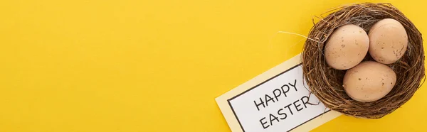 Vista superior de la tarjeta de felicitación con letras de Pascua feliz cerca de huevos de pollo en el nido sobre fondo amarillo colorido, tiro panorámico - foto de stock