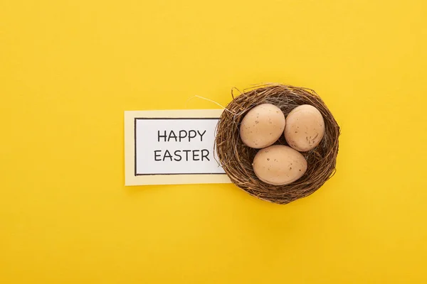 Top view of greeting card with happy Easter lettering near chicken eggs in nest on yellow colorful background — Stock Photo