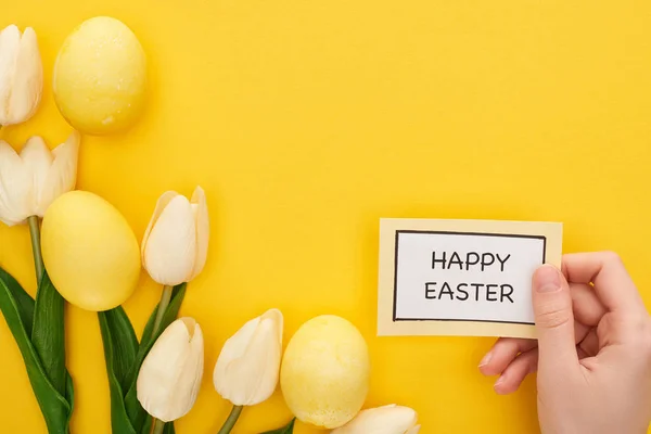 Zugeschnittene Ansicht einer Frau, die eine Karte mit fröhlichem Osteraufdruck in der Nähe von Tulpen und bemalten Ostereiern auf buntem gelben Hintergrund hält — Stockfoto