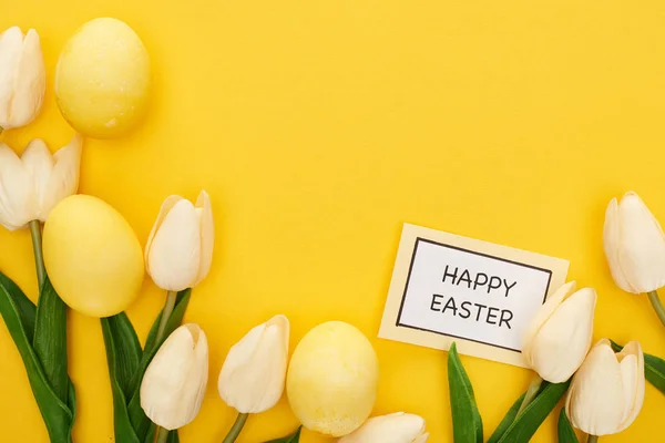 Top view of tulips and painted Easter eggs near card with happy Easter lettering on colorful yellow background — Stock Photo