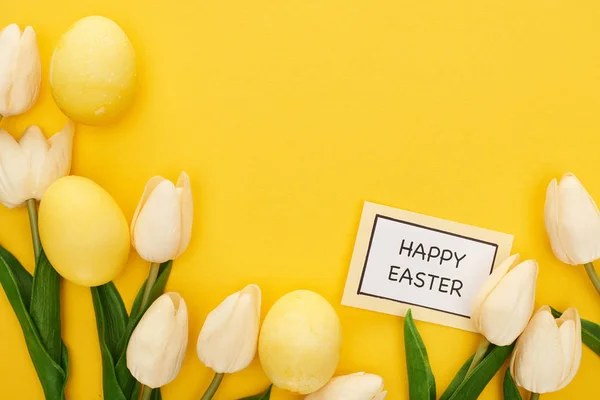 Vista superior de tulipanes y huevos de Pascua pintados cerca de la tarjeta con letras de Pascua feliz sobre fondo amarillo colorido - foto de stock