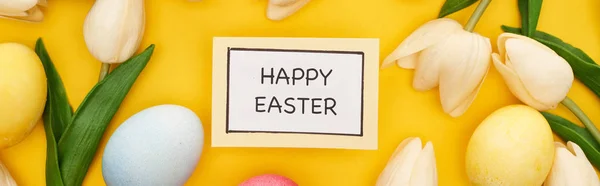 Top view of tulips and painted Easter eggs around card with happy Easter lettering on colorful yellow background, panoramic shot — Stock Photo