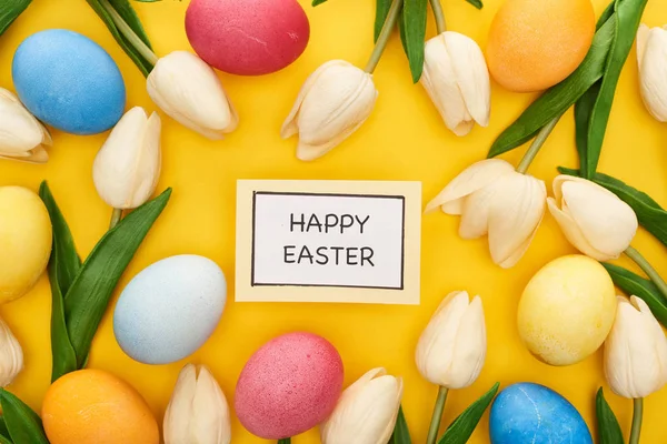 Top view of tulips and painted Easter eggs around card with happy Easter lettering on colorful yellow background — Stock Photo