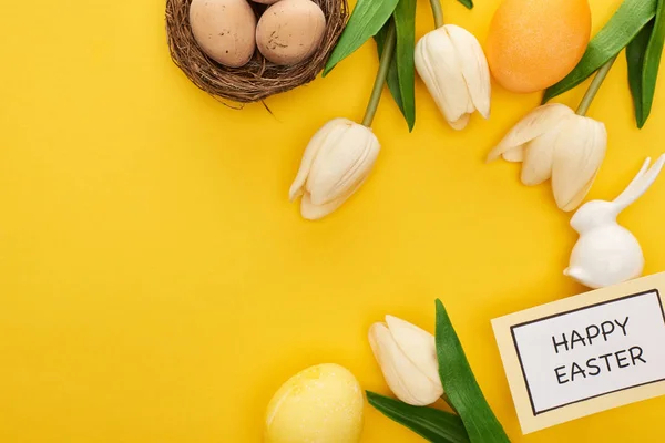 Draufsicht auf Tulpen, Grußkarte mit fröhlichem Osteraufdruck, Osterhase und Eier im Nest auf gelbem, buntem Hintergrund — Stockfoto