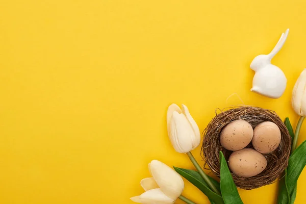 Vue du dessus des tulipes et des œufs de poulet dans le nid près du lapin de Pâques sur fond jaune coloré — Photo de stock