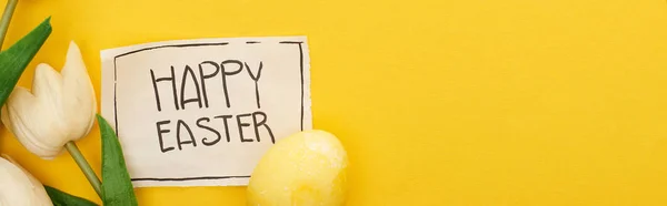 Top view of Easter egg, tulips and greeting card with happy Easter lettering on yellow colorful background, panoramic shot — Stock Photo