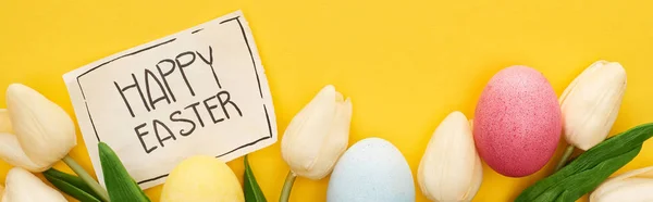 Vista superior de los huevos de Pascua, tulipanes y tarjeta de felicitación con letras de Pascua feliz sobre fondo amarillo colorido, tiro panorámico - foto de stock