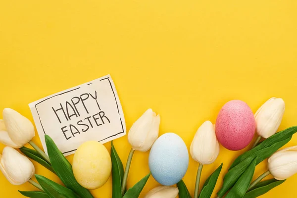 Top view of Easter eggs, tulips and greeting card with happy Easter lettering on yellow colorful background — Stock Photo