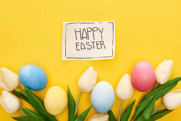 Top view of Easter eggs, tulips and greeting card with happy Easter lettering on yellow colorful background — Stock Photo