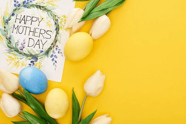 Top view of spring tulips, Easter eggs and happy mothers day greeting card on colorful yellow background — Stock Photo