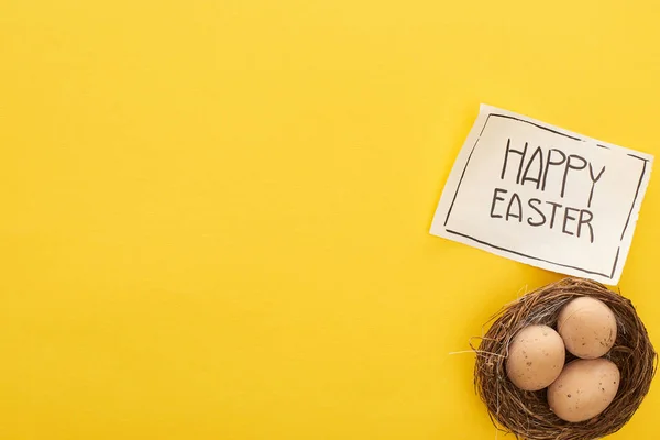 Vista superior de huevos de pollo en nido y tarjeta de felicitación con letras de Pascua feliz sobre fondo amarillo colorido - foto de stock