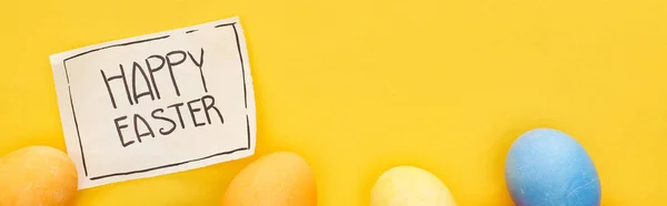 Top view of painted eggs and greeting card with happy Easter lettering on yellow colorful background, panoramic shot — Stock Photo