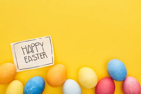 Vue du haut des œufs peints et carte de vœux avec joyeux lettrage de Pâques sur fond jaune coloré — Photo de stock