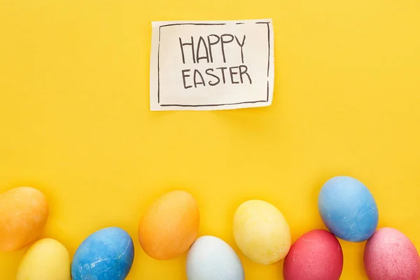 Top view of painted eggs and greeting card with happy Easter lettering on yellow colorful background — Stock Photo