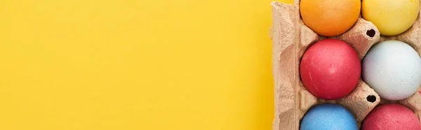 Vue de dessus des oeufs de Pâques peints multicolores dans un récipient en carton sur fond jaune avec espace de copie, vue panoramique — Photo de stock