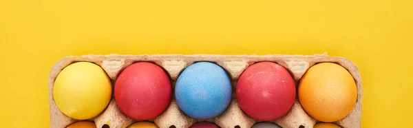 Vista superior de ovos de Páscoa pintados multicoloridos em recipiente de papelão no fundo amarelo, tiro panorâmico — Fotografia de Stock
