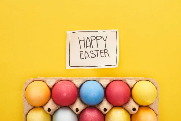 Вид зверху пофарбовані яйця в коробку і вітальна листівка з щасливим Великоднім листям на жовтому барвистому фоні — стокове фото