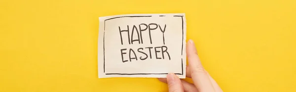 Cropped view of woman holding greeting card with happy Easter lettering on yellow background, panoramic shot — Stock Photo