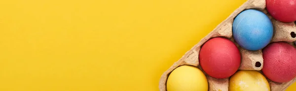 Top view of multicolored painted Easter eggs in cardboard container on yellow background with copy space, panoramic shot — Stock Photo