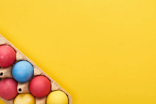 Vista superior de ovos de Páscoa pintados multicoloridos em recipiente de papelão no fundo amarelo com espaço de cópia — Fotografia de Stock