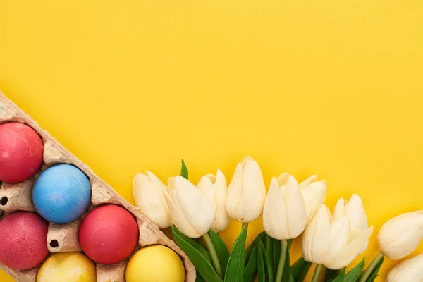 Vista superior de tulipanes y huevos de Pascua pintados multicolores en contenedor de cartón sobre fondo amarillo colorido — Stock Photo