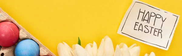 Vue du haut des œufs peints, tulipes et carte de vœux avec joyeux lettrage de Pâques sur fond jaune coloré, vue panoramique — Photo de stock