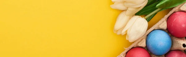 Top view of tulips and multicolored painted Easter eggs in cardboard container on colorful yellow background, panoramic shot — Stock Photo