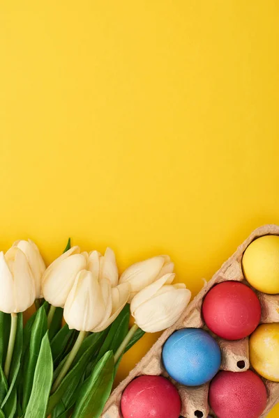 Верхний вид тюльпанов и разноцветных окрашенных пасхальных яиц в картонный контейнер на красочном желтом фоне — стоковое фото