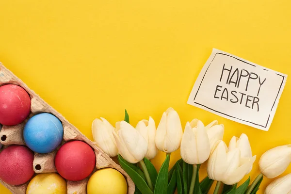 Vista dall'alto di uova dipinte, tulipani e biglietto di auguri con felice iscrizione pasquale su sfondo giallo colorato — Foto stock