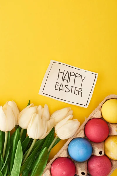 Vista dall'alto di uova dipinte, tulipani e biglietto di auguri con felice iscrizione pasquale su sfondo giallo colorato — Foto stock