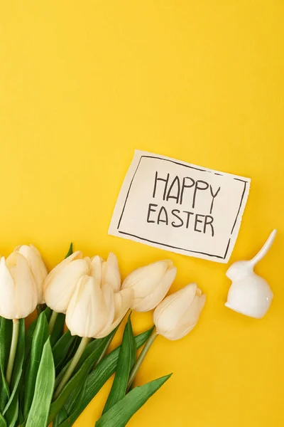 Draufsicht auf Tulpen, Zierhasen und Grußkarte mit fröhlichem Osteraufdruck auf gelbem, buntem Hintergrund — Stockfoto