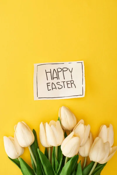 Top view of tulips and greeting card with happy Easter lettering on yellow colorful background — Stock Photo