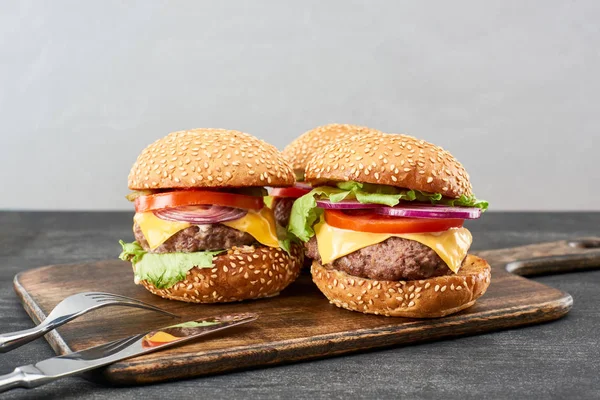 Delicious fresh meat cheeseburgers on wooden board near cutlery — Stock Photo