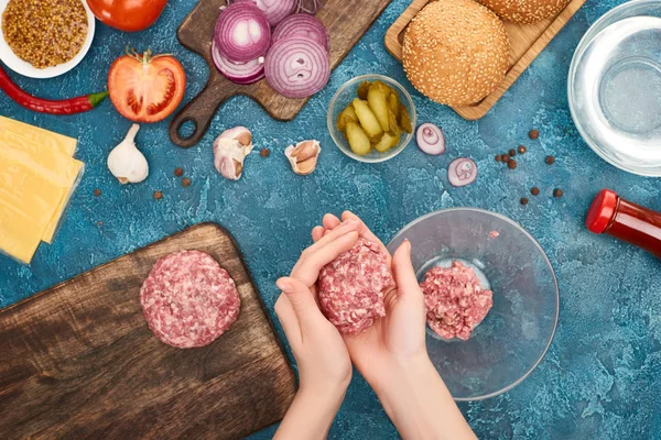 Draufsicht einer Frau, die Schnitzel für frische Burger in der Nähe von Zutaten auf blau strukturierter Oberfläche herstellt — Stockfoto