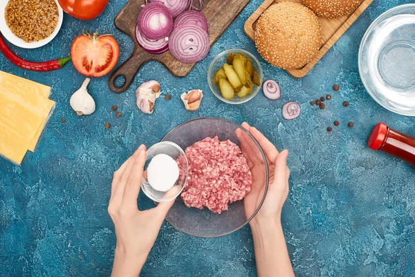 Vista superior de la mujer añadiendo sal a la carne picada cerca de los ingredientes de la hamburguesa en la superficie de textura azul - foto de stock